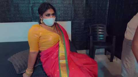 Media: Video of a South Asian woman in a yellow blouse, red sari with gold border, and white mask, sitting on a bed in a dimly lit room with dark curtains and a black chair.