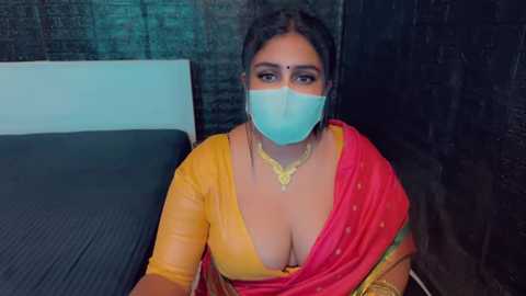 Media: Video of a South Asian woman with medium brown skin, wearing a yellow blouse and red sari with a deep neckline, and a blue face mask, sitting on a bed in a dimly lit room.