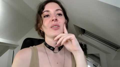 Media: Video of a young Caucasian woman with fair skin, dark brown hair, and brown eyes, wearing a black leather choker and a beige top, touching her cheek, in a modern room with white walls and a ceiling fan.