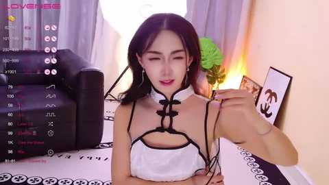 Media: A video of an Asian woman with long black hair, wearing a revealing white dress with black trim, sitting at a table, holding a smartphone.