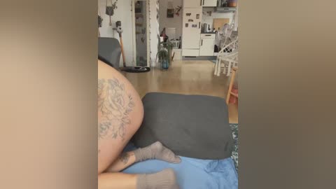 Media: Video of a tattooed woman's bare buttocks and legs, lying on a blue blanket, in a cozy, cluttered living room with wooden floors and white furniture.