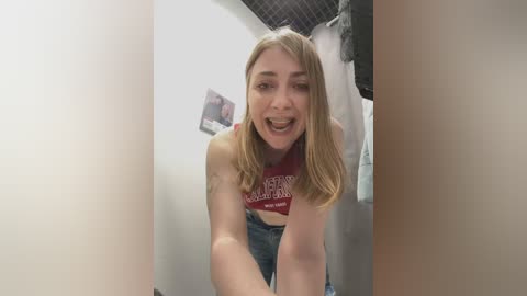 Media: Video of a young Caucasian woman with blonde hair, wearing a red \"GO RUTGERS\" crop top and jeans, smiling in a coin-operated photo booth.