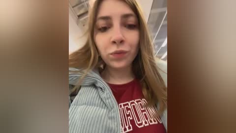 Media: Video of a young woman with long, blonde hair, wearing a red T-shirt with white text and a blue striped hoodie, in an indoor setting with beige walls and visible ceiling beams.