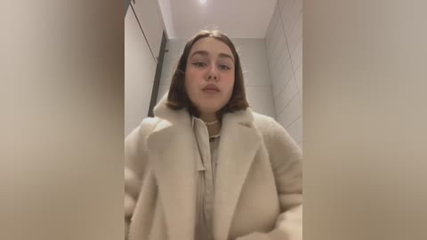 Media: Video of a young woman with fair skin and straight, shoulder-length brown hair, wearing a beige coat, standing in a narrow, tiled hallway.