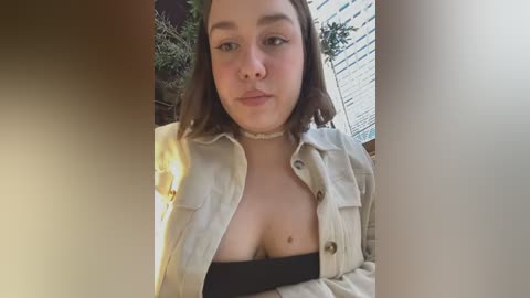 Media: Video of a young woman with light skin, medium-length brown hair, wearing a beige jacket, black top, and choker. She's indoors with a window and greenery visible behind her.