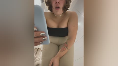 Media: A video captures a young woman with short, curly brown hair, medium build, and light skin, wearing a strapless black top and beige pants, holding a light blue phone, with a visible tattoo on her right arm.