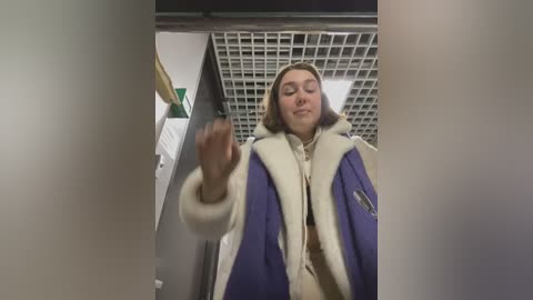 Media: A video shows a young woman with shoulder-length light brown hair, wearing a blue and white fur-trimmed coat, standing in a dimly lit, grid-patterned ceiling hallway.