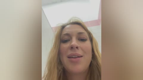 Media: A close-up video of a young Caucasian woman with long blonde hair, wearing minimal makeup and a light pink top. The image is slightly blurred, taken from a low angle, and shows a bathroom setting with white tiles and a pink stripe on the wall.