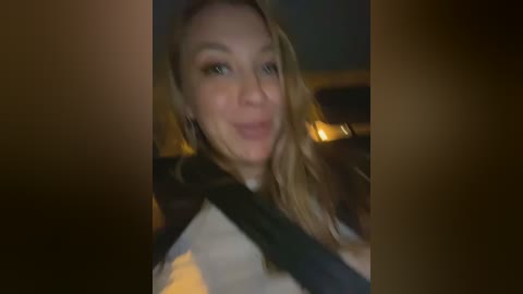 Media: A dimly lit video shows a smiling woman with long blonde hair, wearing a black and white jacket, seated in a car with a seatbelt across her chest. The background is blurred.