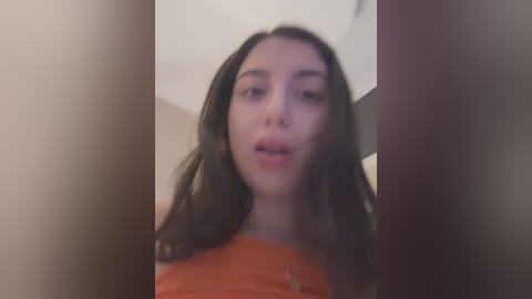 Media: A blurry video of a young woman with long, dark hair, fair skin, and a round face, wearing an orange top, taken indoors with a soft focus.