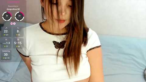 Media: A video of a young woman with long brown hair, wearing a white crop top, standing in a bedroom. The image includes a digital overlay with a \"Deepfake\" watermark.