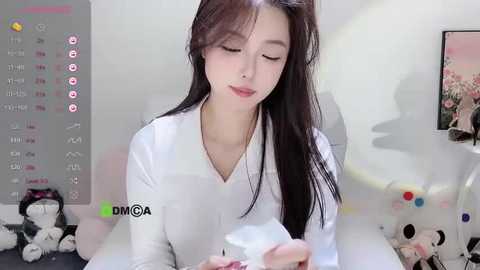 Media: Video of a young East Asian woman with long black hair, wearing a white V-neck shirt, sitting on a bed, holding a white object, in a cluttered bedroom with a calendar and stuffed toys.