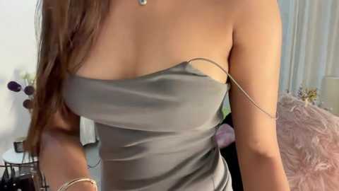 Media: Video of a woman with fair skin, wearing a strapless, grey dress with a thin gold chain. Background features a white wall, a plant, and a pink cushioned sofa.