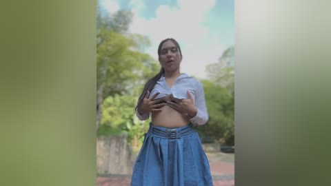 Media: A video of a young, light-skinned woman with long dark hair in a ponytail, wearing a white button-up shirt lifted to reveal her breasts, and a high-waisted denim skirt. She stands in a green-tinted garden, partially obscured by foliage.