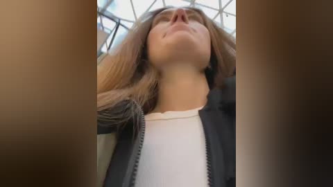 Video of a Caucasian woman with long, straight blonde hair, wearing a white shirt under a dark jacket, looking up with a serene expression, surrounded by large, brown-toned window frames.