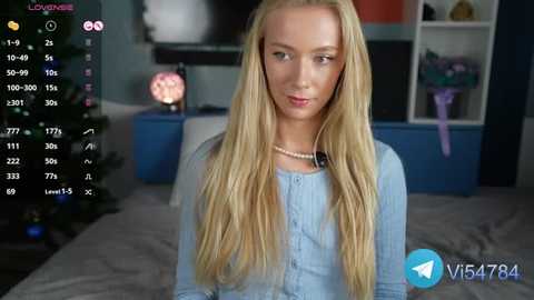 Media: Video of a blonde woman with long hair in a light blue shirt, sitting in a dimly lit room with a Christmas tree and shelves in the background.