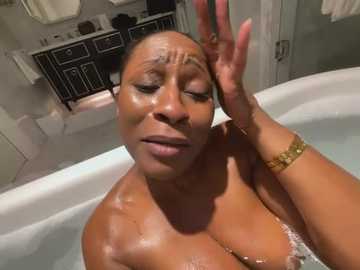 Media: Video of a Black woman with short hair, wet and glistening, sitting in a white bathtub, hand on her forehead, in a modern bathroom with white tiles and black cabinets.