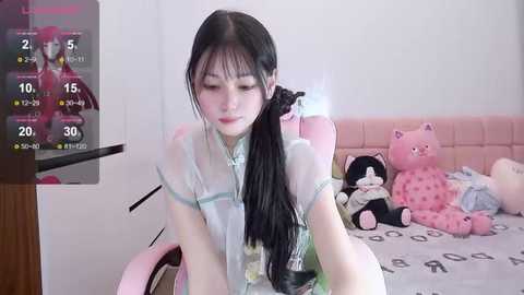 Media: A video of an East Asian woman with long black hair, wearing a light green blouse, playing a video game on a pink gaming chair. A pink teddy bear is beside her, and a TV screen displays game stats.