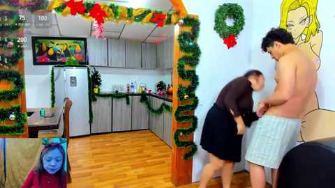 Media: Video of a festive kitchen with green garlands, wooden floors, and a framed anime character; a shirtless man in a towel and a woman in a black dress adjusting his shorts.