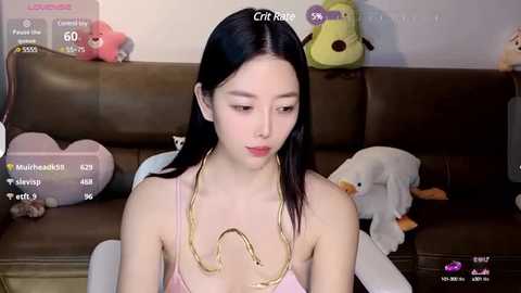 Media: Video of a young Asian woman with fair skin, dark hair, wearing a pink spaghetti-strap top, gold necklace, and seated on a brown leather couch. Background includes plush toys and a white wall.