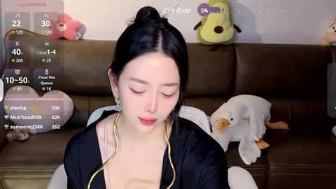 Media: A video of a young Asian woman with fair skin, black hair in a bun, wearing a black jacket over a beige top, sitting on a brown leather couch with plush toys, including a cat and guitar, in a cozy living room.