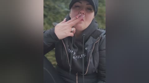 Media: A video of a young person with light skin, wearing a dark hoodie and hat, making a shushing gesture outdoors. The blurred background features greenery.