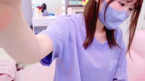 Media: Video of a young Asian woman with straight brown hair, wearing a light blue surgical mask and gown, taking a selfie in a bright, sterile medical environment.