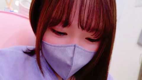 Media: Video of a young Asian woman with straight, shoulder-length brown hair and bangs. She wears a light blue surgical mask and a light blue gown, eyes closed, possibly resting or in a contemplative state. Background is blurred.