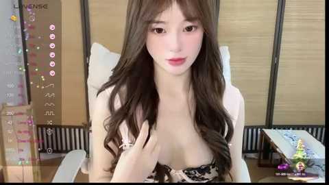 Media: A video of a young Asian woman with long, wavy brown hair, fair skin, and small breasts, wearing a black and white patterned bra, sitting at a desk. Background includes a white chair and a beige wall with a grid pattern.