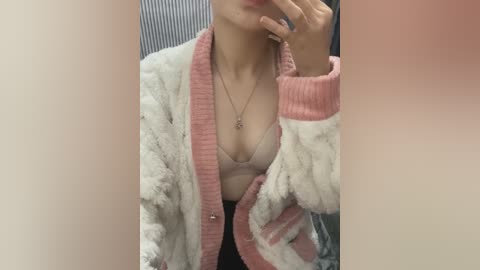 Media: A video of a woman in a pink, fluffy cardigan over a beige bra, partially visible. Her face is partially obscured, holding a phone. Background shows a blurred, striped wall.