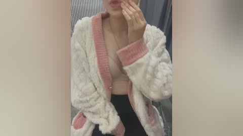 Media: A video of a fair-skinned woman with light brown hair, wearing a fluffy, pink-trimmed white robe, partially unbuttoned, revealing a pink bra. She is indoors, possibly in a changing room, with a blurred background.