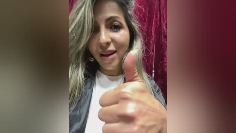 Media: Video of a woman with shoulder-length blonde hair, medium skin tone, giving a thumbs-up gesture in a red velvet background. She's wearing a dark jacket over a white shirt.