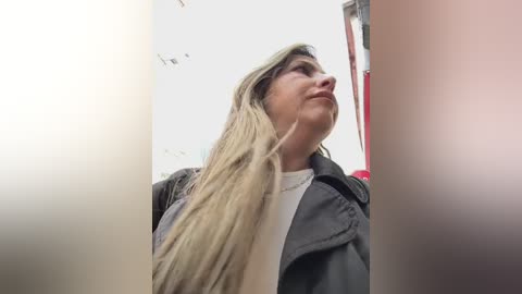 Media: Video of a woman with long blonde hair, wearing a gray jacket over a white shirt, looking upward in a reflective surface, likely a mirror.