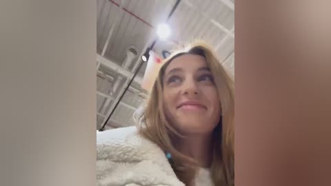 Media: Video of a smiling young woman with long blonde hair, wearing a fuzzy white hat and a textured white sweater, standing in a brightly lit, white room with visible ventilation ducts.