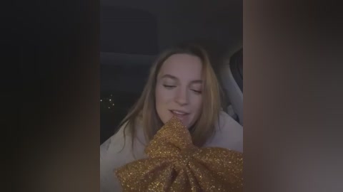 Media: Video of a young woman with fair skin and long, straight blonde hair, wearing a white top, eating a glittery gold bow inside a dimly lit car.