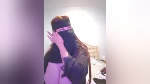 Media: Video of a person with long red hair, wearing a black veil, holding a hand over their face, standing in a dimly lit bedroom with a bed and clothes visible in the background.