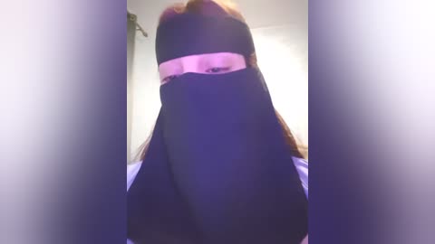 Media: A video of a person wearing a black niqab with a pink strip over their eyes, standing in a softly lit room with a blurred background.