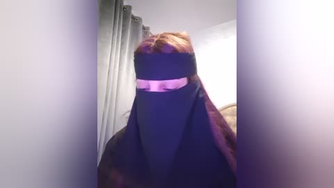 Media: Video of a person wearing a black niqab with a pink strip over the eyes, standing in a room with gray curtains and white walls.