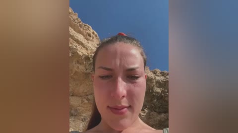 Media: Video of a young woman with light skin and brown hair tied back, standing against a rocky cliff under a clear blue sky. Her expression is neutral, and she wears a light-colored top.