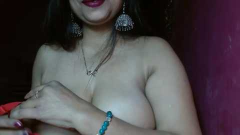 Media: A video of a topless woman with medium skin tone, dark hair, red lipstick, wearing large silver earrings, a necklace, and a blue bracelet. She is indoors against a dark background.