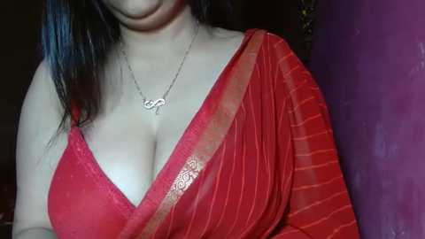 Media: Video of a fair-skinned woman with long black hair, wearing a red saree with gold border, exposing cleavage, and a silver necklace. Background features a purple wall and indistinct objects.