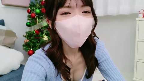 Media: Video of an East Asian woman with long brown hair, wearing a white face mask and light blue knitted sweater, standing in a festive room with a decorated Christmas tree and white curtains.