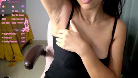 Media: Video of an Asian woman with straight black hair, light skin, and small breasts, wearing a black spaghetti-strap dress, raising her arm to show her armpit hair, in a room with a yellow floral tablecloth, a computer monitor, and a brown chair.