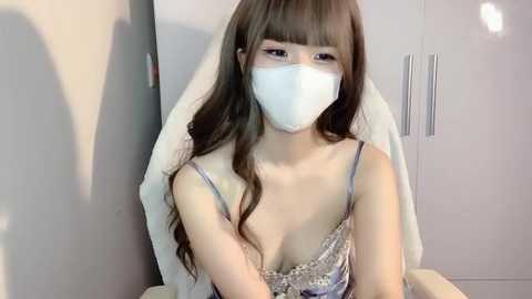 Media: Video of a young Asian woman with long brown hair, wearing a white face mask, a light blue camisole, and a white towel draped over her shoulders, seated in a modern, minimalist room with white cabinets and silver handles.