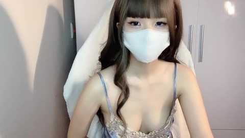 Media: Video of an Asian woman with long brown hair, wearing a white surgical mask, a lace-trimmed, low-cut blue dress, and a white towel draped around her shoulders. The background features a plain, light-colored wall.