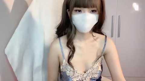 Media: Video of an Asian woman with long brown hair, wearing a light blue face mask and a lace-trimmed blue camisole, standing in a white-tiled bathroom with a towel hanging on a rack.