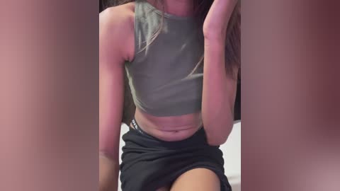 Media: Video of a light-skinned woman with long brown hair, wearing a tight, sleeveless grey crop top and a black mini skirt, sitting with legs apart, partially obscured by blurry figures in the foreground.