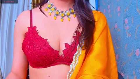 Media: Video of a South Asian woman with light skin, wearing a red lace bra and yellow sari, adorned with a colorful necklace, against a textured blue wall.