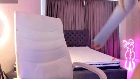 Media: A video of a modern bedroom featuring a gray upholstered bed with a tufted headboard, white bedding, and a minimalist nightstand with a glowing neon sign. The room has dark gray curtains and soft lighting.