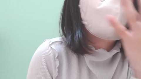 Media: Video of a woman with short black hair wearing a white mask and a light gray, ribbed-textured blouse, her face partially obscured by the mask, against a pastel green background.